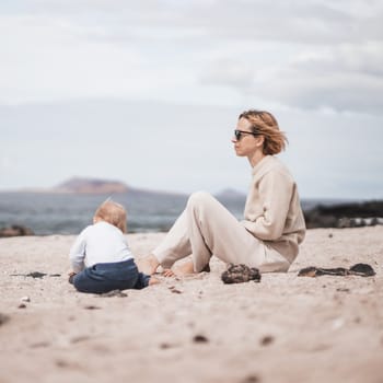 Mother enjoying winter beach vacations playing with his infant baby boy son on wild volcanic sandy beach on Lanzarote island, Canary Islands, Spain. Family travel and vacations concept.