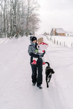 Smiling mother with a little girl in her arms walks with her dog along a snowy village road. High quality illustration