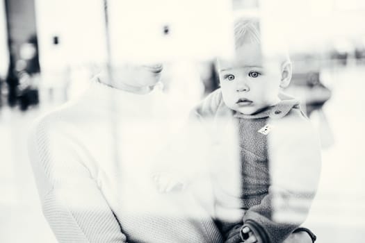 Thoughtful young mother looking trough window holding his infant baby boy child while waiting to board an airplane at airport terminal departure gates. Travel with baby concept