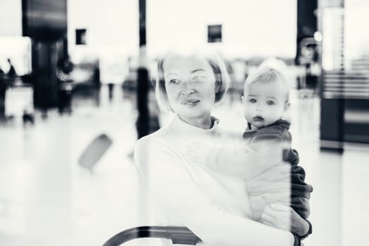 Thoughtful young mother looking trough window holding his infant baby boy child while waiting to board an airplane at airport terminal departure gates. Travel with baby concept