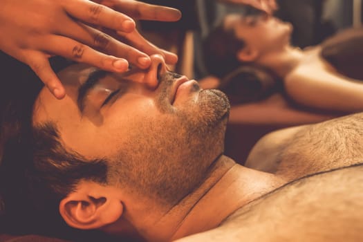 Couple customer enjoying relaxing anti-stress head massage and pampering facial beauty skin recreation leisure in warm candle lighting ambient salon spa in luxury resort or hotel. Quiescent