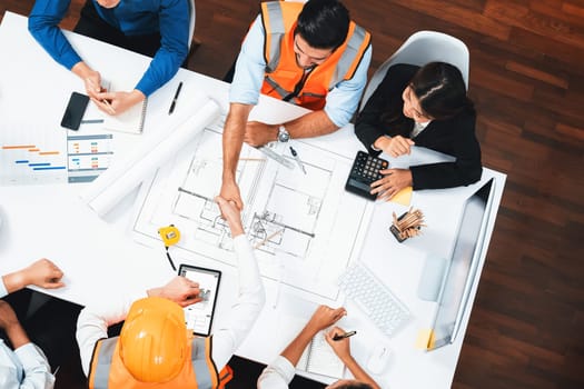 Top panorama of diverse group of civil engineer and client shake hand after make successful agreement on architectural project, review construction plan and building blueprint at meeting table.Prudent