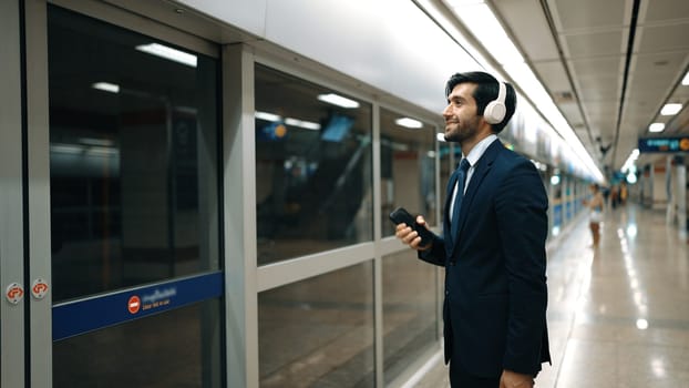 Project manager wearing headphone at train station while holding mobile phone for choosing song. Smart business man listening relaxing music while waiting for train with blurred background. Exultant.