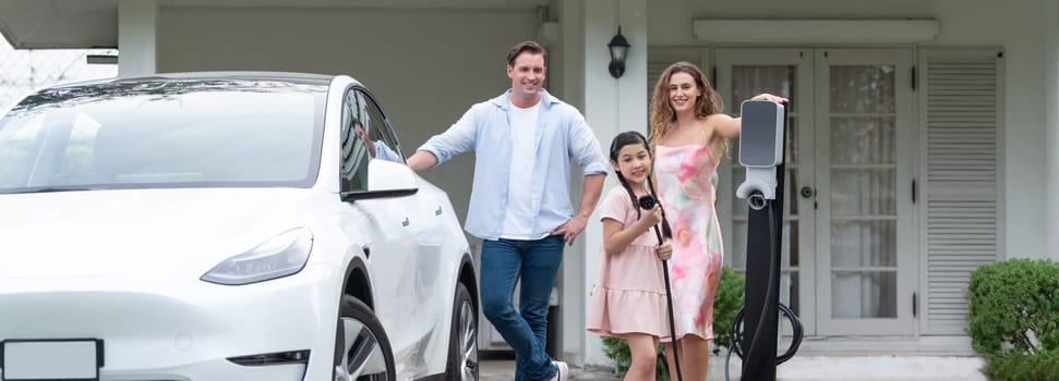 Modern family with environmental conscious recharging EV car with home EV charging station, Happy family with little young girl holding and pointing EV charger at camera. Panorama Synchronos