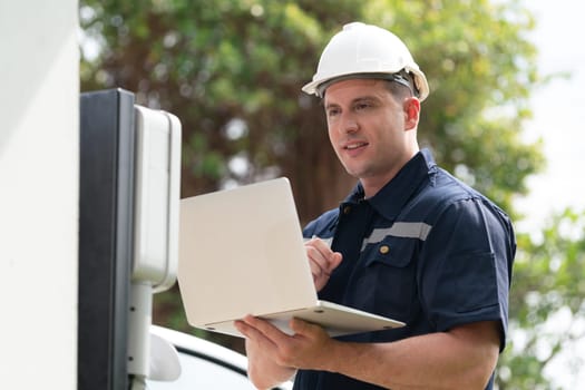 Qualified technician working on home EV charging station installation, making troubleshooting and configuration setup on charging system with laptop for EV at home. Synchronos