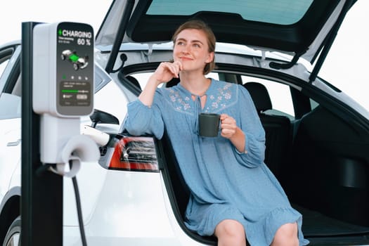 Holiday road trip vacation with young woman sitting in the car trunk with coffee while recharging electric vehicle with alternative energy. Environmental friendly travel wit EV car. Perpetual