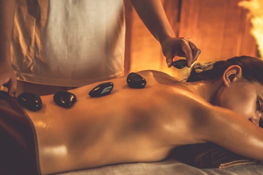 Hot stone massage at spa salon in luxury resort with warm candle light, blissful woman customer enjoying spa basalt stone massage glide over body with soothing warmth. Quiescent