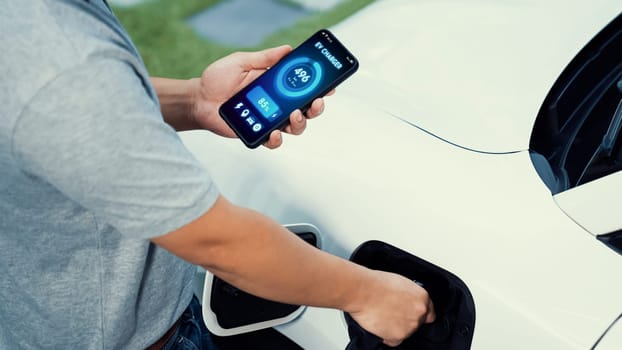 Hand insert EV charger to electric car to recharge battery while check battery status display on smartphone. Future sustainable and clean energy for EV car. Peruse