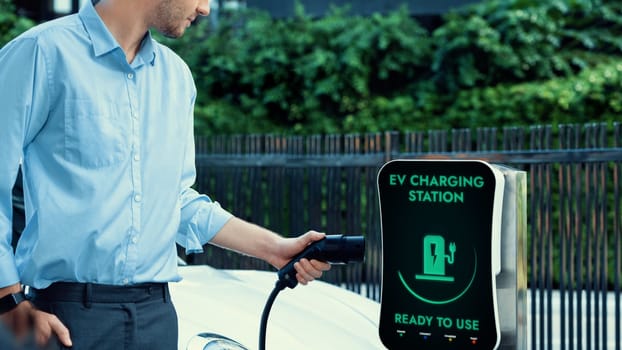 Businessman pull and hold EV charger plug form electric car charging station at city car park area background. Futuristic clean sustainable energy and EV car technological advancement.Peruse