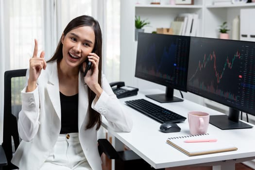 Young beautiful Asian businesswoman talking to broker on phone with looking on pc screen to analyze increased stock exchange market graph at modern office, investing high profit in wealth. Stratagem.