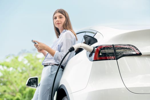 Young woman using smartphone online banking application to pay for electric car battery charging from EV charging station during vacation holiday road trip at national park or summer forest. Exalt