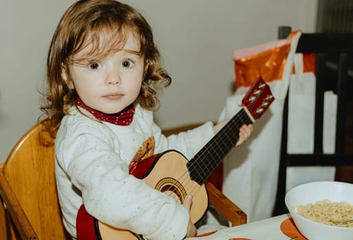 One beautiful Caucasian baby girl with large expressive and brown eyes plays the guitar while sitting at the table in the kitchen, close-up side view.