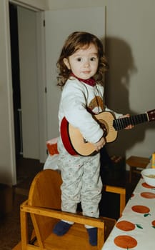One beautiful Caucasian baby girl with large expressive and brown eyes plays the guitar while standing on a wooden chair at the table in the kitchen, close-up side view.