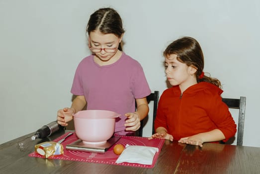 Two beautiful Caucasian brunette girls stand at a table with food and accessories for baking cookies, standing at the table during the day in the kitchen, close-up side view. Step-by-step instructions. Step 1.