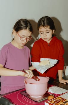 Two beautiful Caucasian brunette girls are standing at a table with food and one is cutting a piece of butter into a bowl for baking cookies, standing at the table in the kitchen during the day, close-up side view. Step-by-step instructions. Step 2.