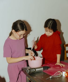 Two beautiful Caucasian brunette girls are standing at the table, and one is pouring the prepared flour mixture into a bowl for baking cookies, and the other is mixing the dough with her hands, close-up side view. Step by step instructions. Step 6.