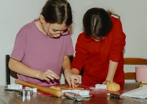 Two beautiful Caucasian brunette girls stand at the table and together remove with their hands the excess cut out yellow shortbread dough lying on a pink silicone mat, close-up side view. Step by step instructions. Step 17