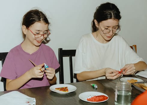 Two beautiful Caucasian happy brunette girls are sitting at the table and decorating baked cookies with paints and chocolate using paints and a tube, close-up side view. Step by step instructions. Step 24