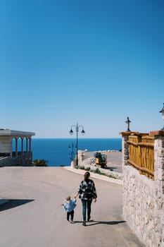 Mom and a little girl walk holding hands along the road overlooking the sea. Back view. High quality photo