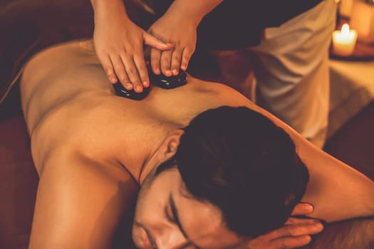 Hot stone massage at spa salon in luxury resort with warm candle light, blissful man customer enjoying spa basalt stone massage glide over body with soothing warmth. Quiescent