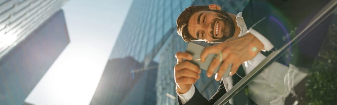 Bottom view of handsome entrepreneur using mobile phone standing on background of skyscrapers