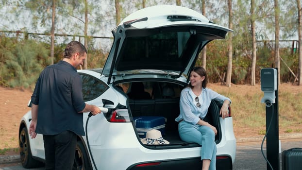 Couple recharge their EV car with electric battery charging station at rest stop during their road trip travel during autumnal season to national park. Relaxing holiday with eco friendly car. Exalt