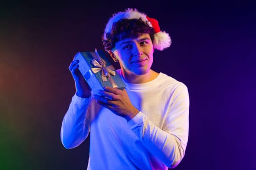 Excited man in Santa hat hold gift box with bow. He is happy and flattered by attention. Guy smiling with present on neon background. Christmas time. High quality photo