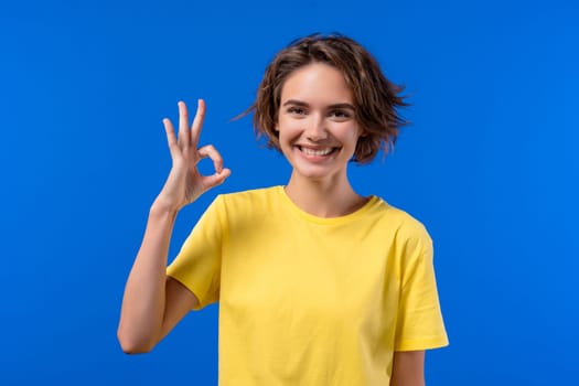 Pretty woman makes hand sign okay, ok gesture. Happy girl, correct perfect choice, great deal, blue background. Positive female model smiles to camera, approval, trust concept. quality