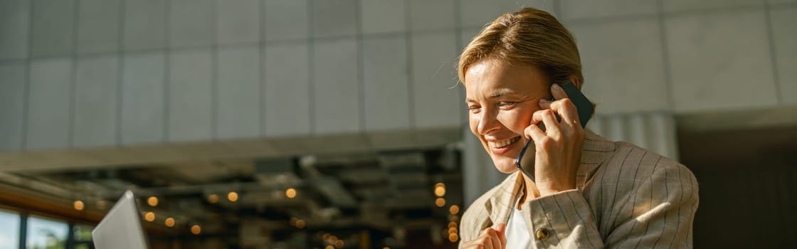 Woman manager talking on phone with client while working on laptop in modern coworking