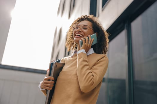 Smiling woman manager is talking phone while standing with laptop on modern building background