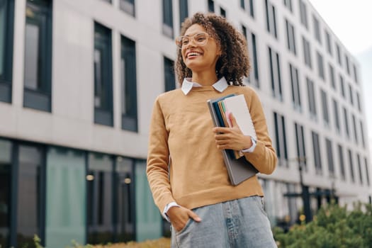 Young smiling female student standing with laptop and note pads on modern building backgrounds