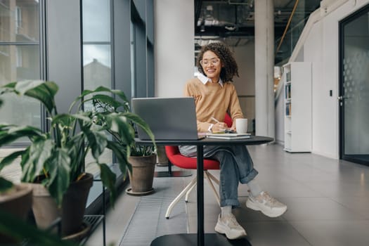 Business woman in eyeglasses working on laptop and making notes while sitting in modern office