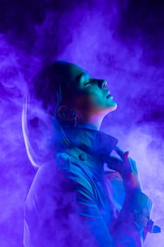 Young woman in neon multi-colors light on smoke, steaming background. Millennial enigmatic futuristic lady in fashion trench coat, stylish outfit