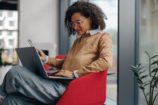 Stylish business woman is use phone while working on laptop in modern office near window