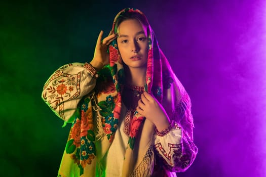 Modern young woman in traditional ukrainian handkerchief, necklace and embroidered blouse at multi-colors neon light smoke background. Ukraine, style, folk, culture. High quality photo