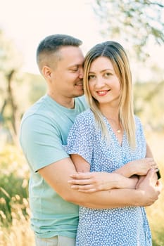 Guy hugs smiling girl from behind, touching her cheek with his nose while standing in the garden. High quality photo