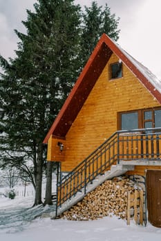 Two-story wooden cottage with a woodpile under the steps near a spruce tree under snowfall. High quality photo