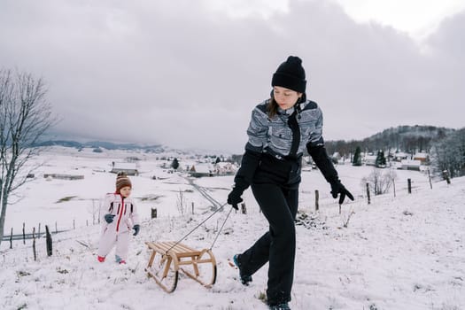 Mom is pulling a sled on a rope up the mountain, looking back at the little girl following behind her. High quality photo