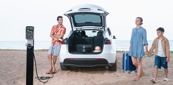 Family vacation trip traveling by the beach with electric car, lovely family taking luggage out while charging EV car battery with clean energy. Alternative family travel by eco-friendly car.Perpetual