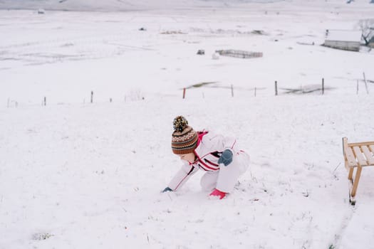 Little girl makes a snowball while squatting on a snowy clearing near a wooden sleigh. High quality photo
