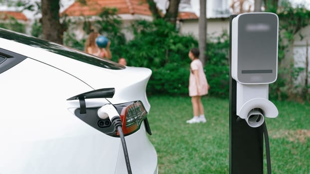 Electric vehicle recharge from home charging station on background of happy and playful family playing together. EV car using alternative and sustainable energy for better future Synchronos