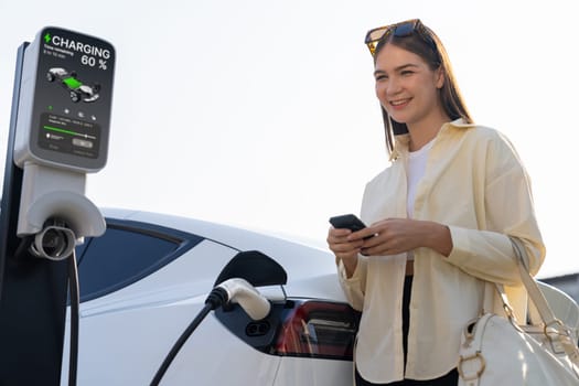 Young woman holding shopping bag and use smartphone to pay for electricity for recharging EV car battery from charging station at city mall parking lot. Modern woman go shopping by eco car. Expedient