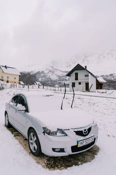 Snow-covered car with raised windshield wipers stands near a cottage. High quality photo