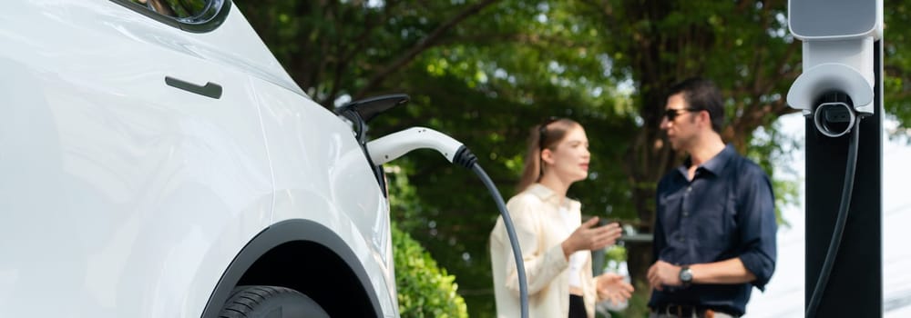 Young couple recharge electric car battery from charging station in green city park in springtime. Rechargeable EV car for sustainable environmental friendly urban travel lifestyle. Panorama Expedient