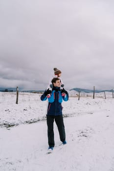 Dad with a little girl on his shoulders walks along a fenced snowy pasture looking to the side. High quality photo