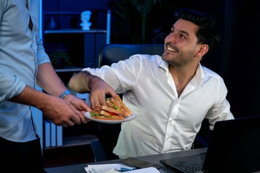 Coworker partners with happy smiling face sharing delicious sandwich dish with friend at night time. Hungry creative colleagues enjoy eating fast food at workplace with neon blue light. Sellable.