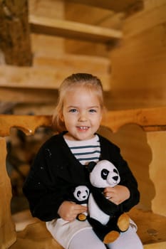 Little smiling girl sitting with soft toy pandas on a wooden chair. High quality photo