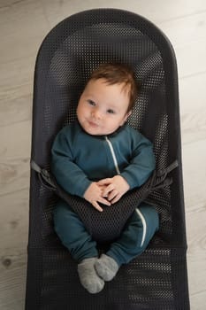 Cute newborn baby dressed in blue overalls sitting in a baby lounger. Vertical photo