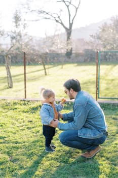 Little girl shows an apple to her dad, who is squatting on the lawn. Side view. High quality photo