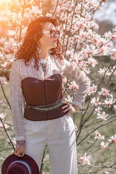 Magnolia park woman. Stylish woman in a hat stands near the magnolia bush in the park. Dressed in white corset pants and posing for the camera